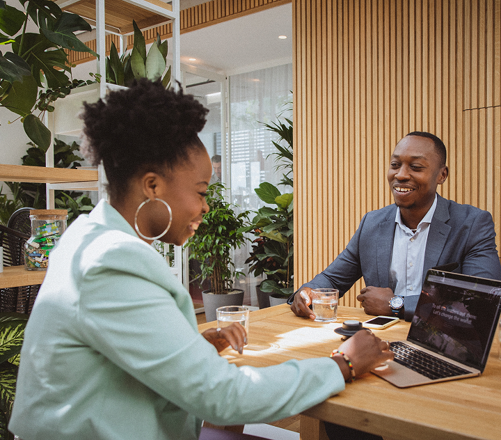 Zwei Personen sitzen im Office an einem Tisch und interagieren miteinander. Die männliche Person hält ein Wasserglas in der Hand und lächelt die weibliche Person an, die in einen Laptop schaut und auf der Tastatur tippt.
