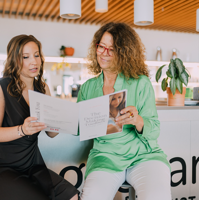 Zwei Personen sitzen im Office an der Küchentheke und zeigen sich gegenseitig Broschüren mit nicht lesbaren Inhalt darauf.
